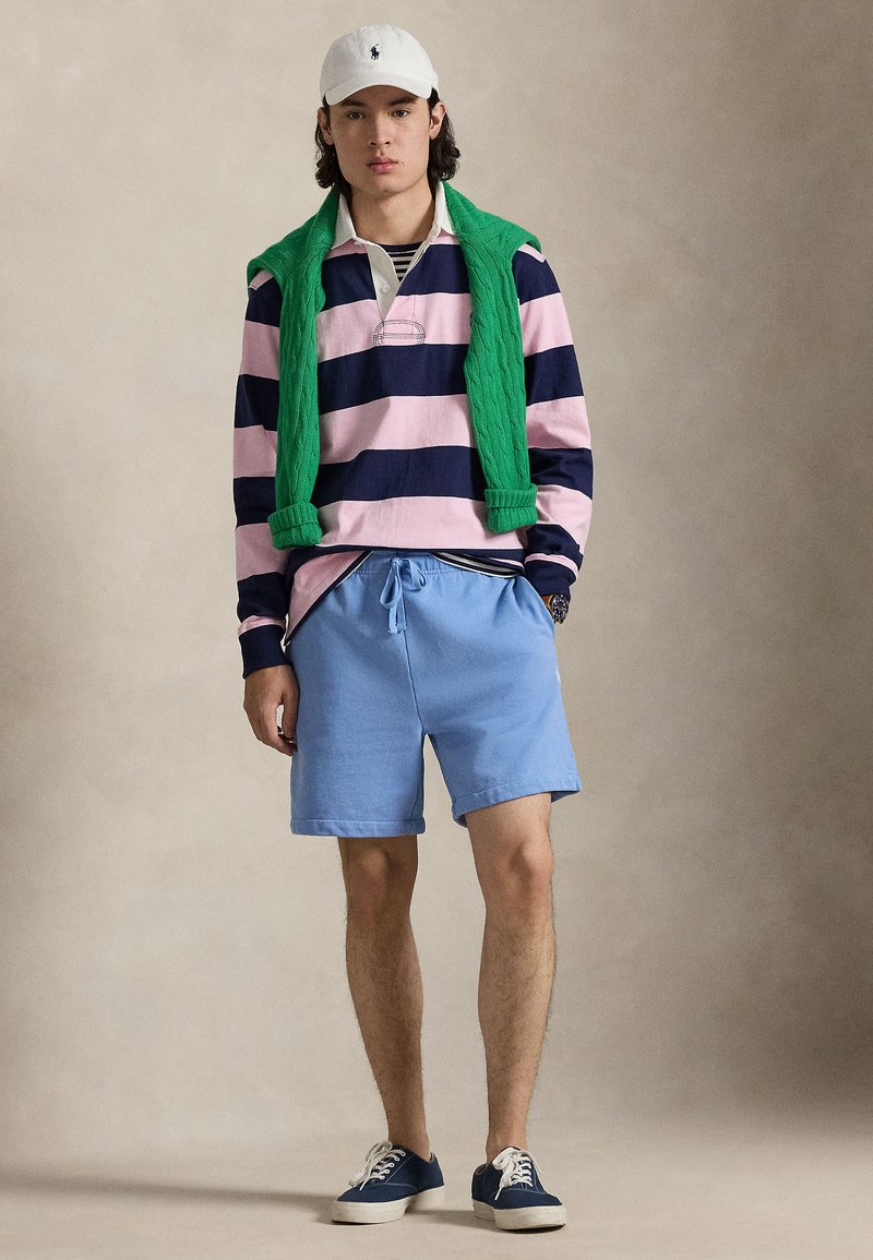 Young man standing wearing white cap, green sweater draped over shoulders, navy and pink striped rugby shirt, light blue shorts, and navy sneakers.