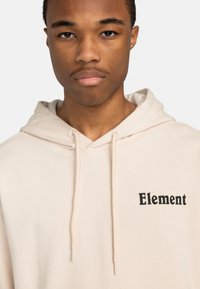 Sweat à capuche beige en matériau doux, avec une poche avant, un cordon de serrage à la capuche et un logo noir "Element" sur la poitrine gauche.