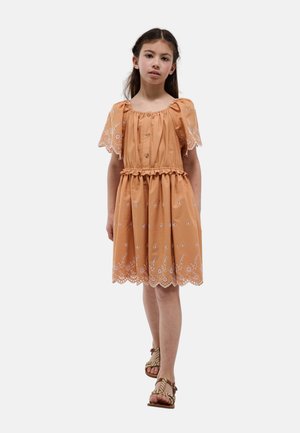 Jeune fille marchant dans une robe marron clair avec broderie florale et manches courtes bouffantes, portant des sandales tressées beiges.