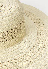Sombrero de paja de ala ancha, de un beige claro, con un diseño texturizado en capas y detalles trenzados a lo largo de la corona y el borde de la ala.