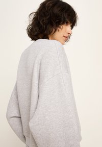 Person mit kurzen, lockigen, dunklen Haaren, die einen lockeren, hellgrauen Sweatshirt trägt und seitlich vor einem schlichten, hellen Hintergrund steht.