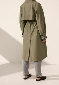 Olijfgroene trenchcoat met een cropped ontwerp, een ceintuur in de taille en zijsplitsen. Draag het over grijze jeans en zwarte instap schoenen.