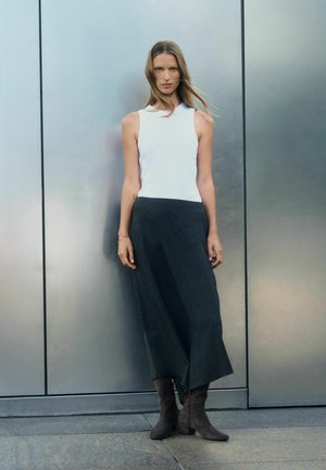 Woman with long hair stands against metal wall wearing white sleeveless top, black wide pants, and dark ankle boots.