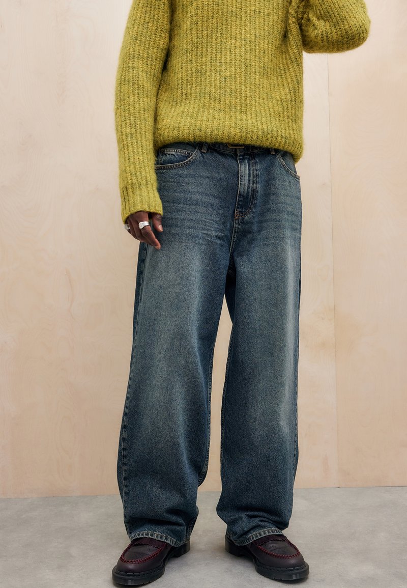 Jeans azuis desgastados e oversized, feitos de denim, com corte direito; combinados com um suéter texturizado verde-oliva e sapatos escuros.