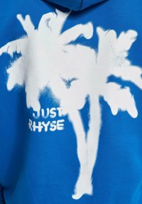 Sudadera azul con un dise�ño de pintura en spray de una palmera blanca y el texto "JUST RHYSE" debajo. Textura suave, estilo casual y colores llamativos.