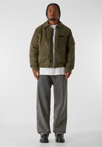 Olivgrön bomberjacka med dragkedja framtill, ribbade manschetter och en foderficka på ärmen, bärs över en vit skjorta och grå jeans.