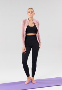 Tenue de sport comprenant une veste rose à manches longues, un crop top noir, et un legging noir taille haute ; posée sur un tapis de yoga violet.