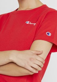 Rød bomulds t-shirt med rund hals og korte ærmer, med et lille broderet logo på brystet og ærmet.