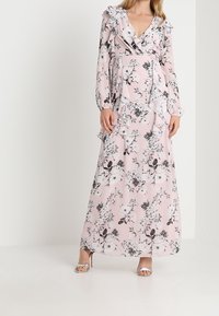 Abito lungo rosa con motivo floreale nero. Presenta maniche lunghe, volants lungo il colletto e in vita, e un design avvolgente.