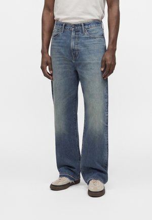 Mand iført løstsiddende blå jeans og beige sneakers står mod en ensfarvet lys baggrund.