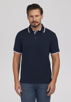 Mężczyzna z brodą i tatuażem nosi granatową koszulkę polo z białym obszyciem oraz niebieskie dżinsy, stoi na tle jednolitego jasnoszarego tła.