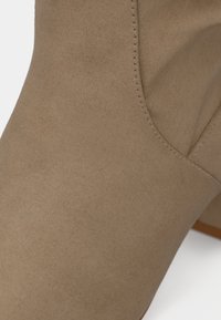 Béžová semišová členková čižma s hladkou textúrou, zaokrúhlenou špičkou a prešitým švom pozdĺž horného okraja. Ploché podrážka s minimálnymi detailmi.