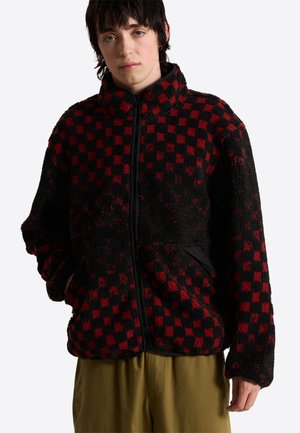 Person trägt eine rot-schwarz karierte Fleecejacke mit Frontreißverschluss und hohem Kragen, steht mit den Händen in den Taschen vor einem weißen Hintergrund.