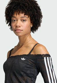 Top negro con escote off-the-shoulder y borde de encaje, con un patrón geométrico y tres rayas blancas en una manga. Logotipo de Adidas en el frente.