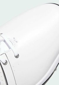 Bílá kožená teniska s plochými bílými tkaničkami, kovovými průchodkami a minimálními švy. Kulatá špička a černá gumová podrážka.