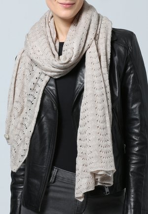 Femme portant une écharpe en tricot beige clair avec un motif ajouré, sur une veste en cuir noire et une chemise noire.