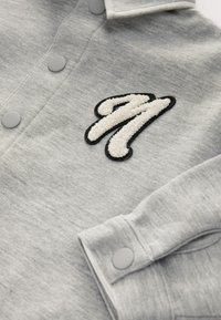 Sweat-shirt gris avec un logo brodé "M" texturé en blanc et noir, des boutons-pression et un détail de revers plié. Texture du tissu douce.