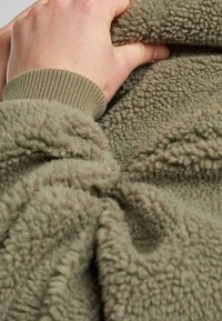 Main tenant un tissu polaire doux, gris verdâtre texturé avec un poignet côtelé visible sur la manche en gros plan.