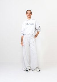 Sivá bavlnená mikina s kapucňou a logom "La Marzia" v modrej farbe, v kombinácii so širokými sivými tepláky. Neformálny dizajn, mäkká textúra a voľný strih.