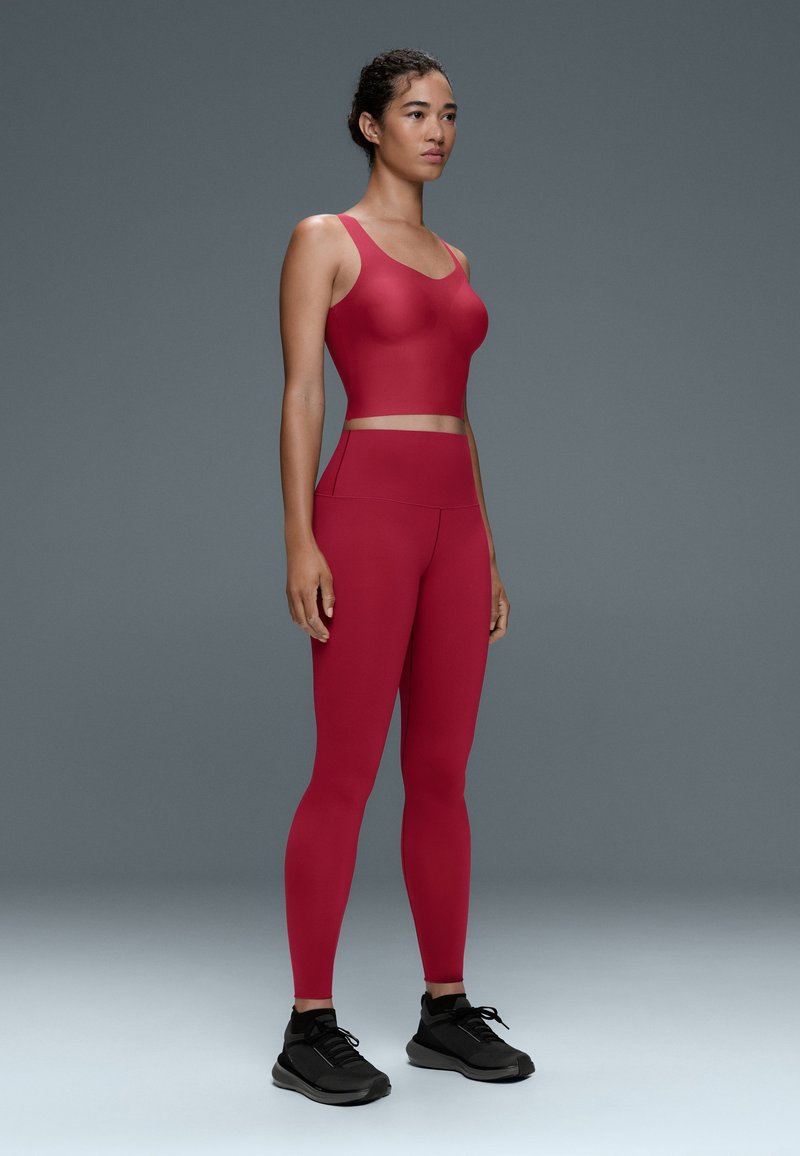 Femme debout portant un crop top rouge ajusté sans manches et des leggings taille haute avec des chaussures de sport noires, sur un fond gris.