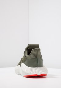 Grön sportskor med en texturerad ovandel, elastisk häl och en vit sula med en röd accent. Förstärkt sömnad bak.