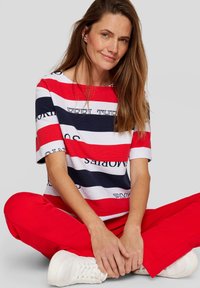 Gestreiftes Kurzarmoberteil in Rot, Weiß und Marineblau mit Schriftzügen; kombiniert mit roten Hosen und weißen Sneakern.