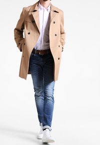 Beige trenchcoat med svarta knappar, bärs över en vit skjorta, i kombination med blå jeans och vita sneakers. Slät tyg, klassisk design.