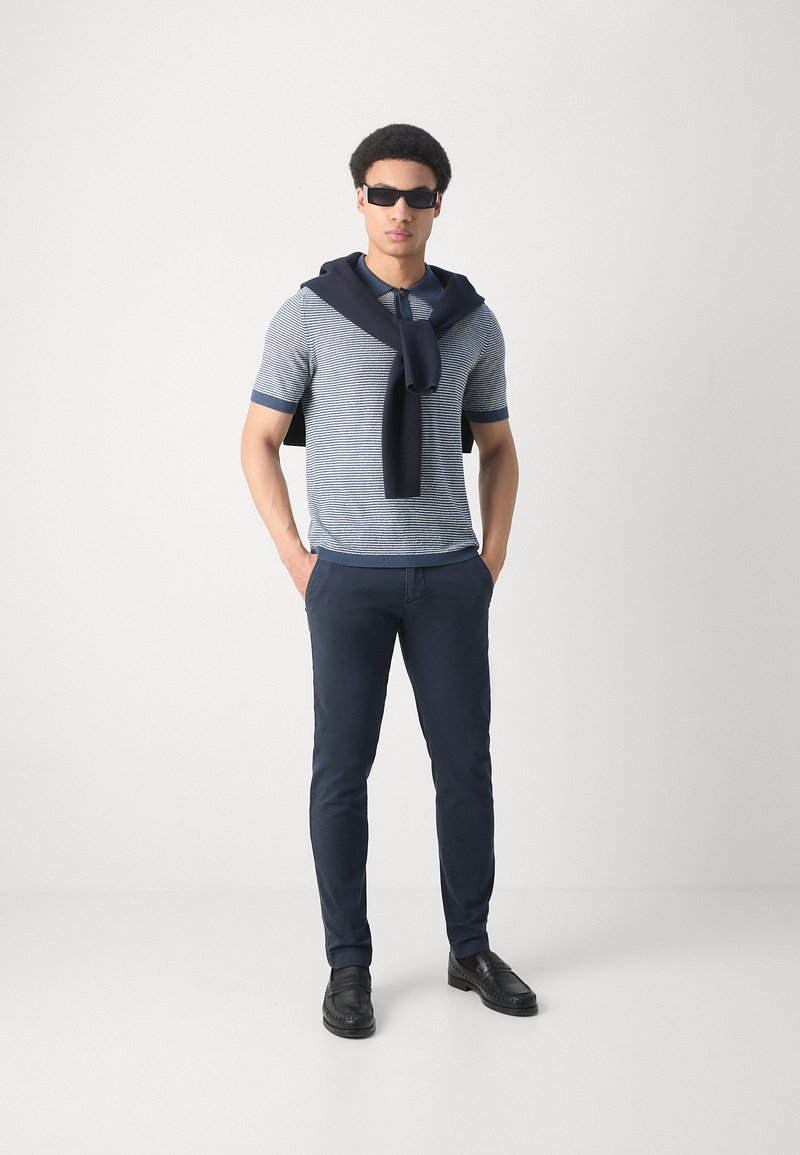 Gestreiftes kurzärmliges Poloshirt in Blau und Weiß, marineblauer Pullover um die Schultern gebunden, marineblaue Hose und schwarze Schuhe. Modell trägt Sonnenbrille.