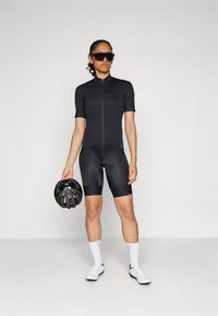 Schwarzes Radtrikot mit kurzen Ärmeln und einem Reißverschluss vorne, kombiniert mit schwarzen Radshorts. Das Model hält einen schwarzen Helm und trägt eine Sonnenbrille.
