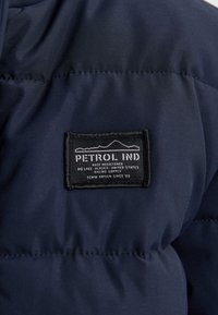 Giacca imbottita blu navy in tessuto con cuciture a sbieco, dotata di un'etichetta nera rettangolare con testo bianco e design grafico di una montagna.