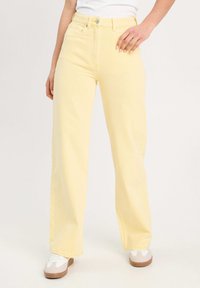 Jean large à taille haute jaune en denim, avec une fermeture à bouton et des poches latérales, associé à des baskets blanches.