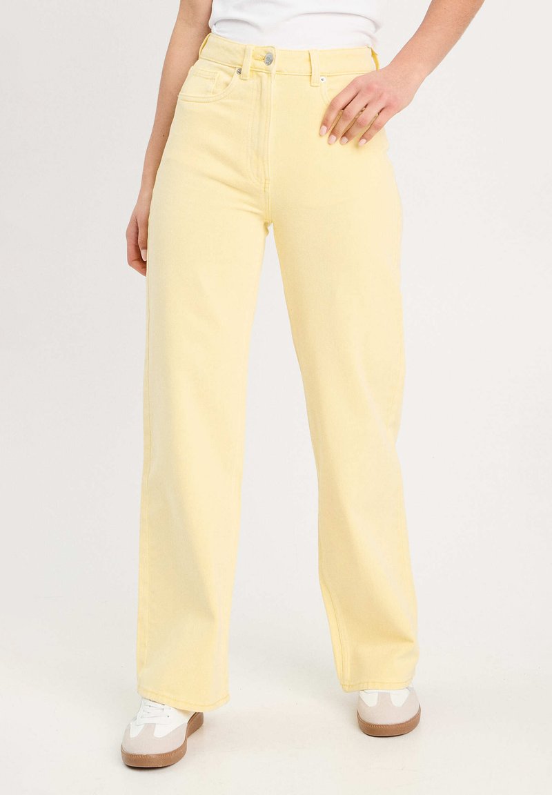 Jean large à taille haute jaune en denim, avec une fermeture à bouton et des poches latérales, associé à des baskets blanches.