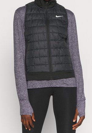 Veste matelassée noire Nike portée par-dessus un haut de sport à manches longues violet avec des trous pour les pouces, associée à un legging noir.
