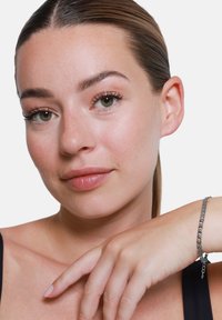 Bracciale d'argento con anelli intrecciati, appoggiato su una mano. Colore della pelle liscio, trucco minimale, sfondo neutro che mette in risalto i tratti del viso.
