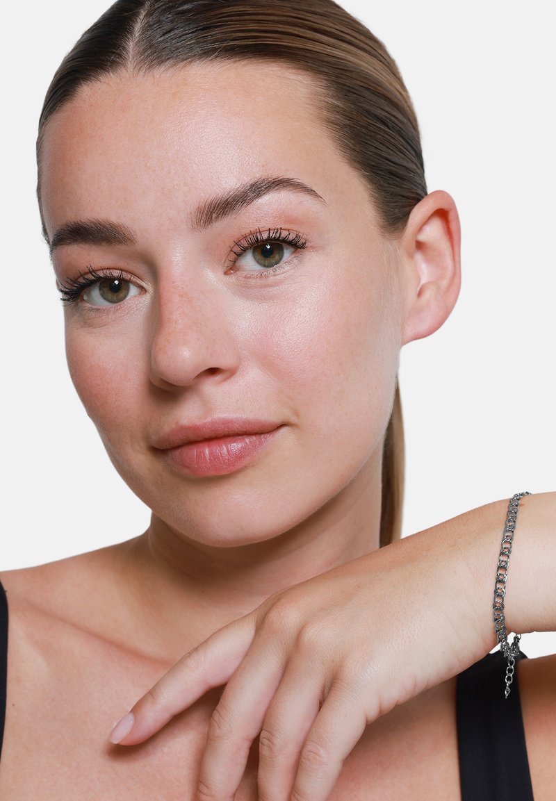 Bracciale d'argento con anelli intrecciati, appoggiato su una mano. Colore della pelle liscio, trucco minimale, sfondo neutro che mette in risalto i tratti del viso.