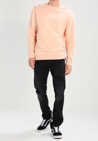 Persona con un sudadera color durazno claro, jeans rasgados negros y zapatillas de lazo en blanco y negro, de pie contra un fondo blanco liso.