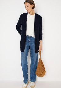 Marinblå lång cardigan med ribbad textur, matchad med en vit tröja och ljusblå jeans. Accessoarer i form av en beige mockaväska och vita sneakers.