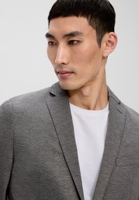 Grau strukturierter Blazer mit einer einzelnen Brusttasche, getragen über einem weißen Rundhals-T-Shirt. Mit Revers und taillierter Passform.
