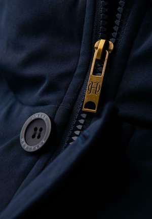 Navytyg i närbild med en guldfärgad metalldragkedja och en rund knapp märkt "Ombre Heritage." Dragkedjan har en texturerad dragtab.