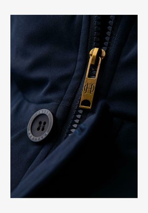 Lähikuva laivastonsinisestä kankaasta, jossa on kultainen metallivetoketju ja pyöreä nappi, jossa lukee "Ombre Heritage". Vetoketjussa on teksturoitu vetotappi.