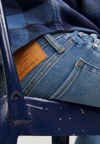 Vaqueros azules de mezclilla con detalles de costura clara, un parche de cuero en la cintura y una camisa azul marino de cuadros parcialmente visible encima.