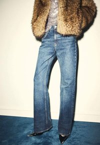 Jeans de mezclilla azul con corte recto, combinados con una chaqueta de piel marrón. Zapatos negros de tacón alto acentúan el conjunto.