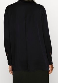 Blouse noire à manches longues en tissu lisse. Présente une coupe droite, des poignets ouverts avec des accents de boutons, et un pli au dos pour plus de confort.