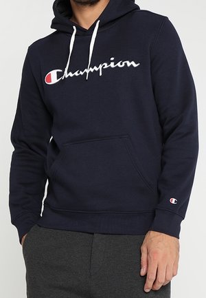 Mężczyzna ubrany w granatową bluzę Champion z białymi troczkami i logo na piersi, w połączeniu z ciemnoszarymi spodniami.