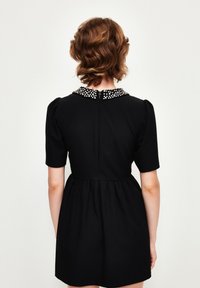 Schwarzes, kurzärmliges Kleid mit tailliertem Schnitt und weit schwingendem Rock. Verfügt über einen strukturierten Kragen, der mit weißen Perlen verziert ist, und einen verdeckten Reißverschluss.