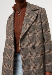 Bruin geruite blazer over een roestkleurige zip-up trui met een ronde metalen ritssluiting, gecombineerd met lichtblauwe jeans.