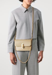Borsa a tracolla in pelle beige con hardware dorato e una tracolla a catena, con chiusura a patta. Il modello indossa un completo a scacchi bianco e nero.