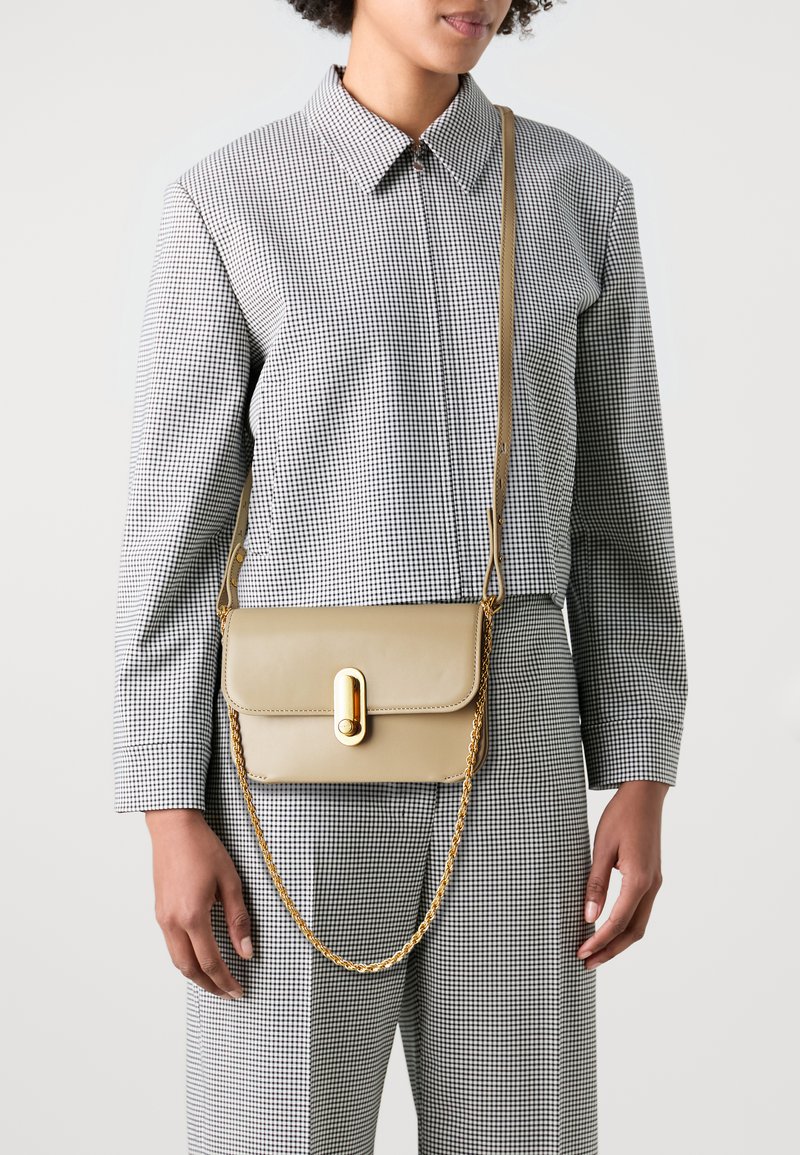 Borsa a tracolla in pelle beige con hardware dorato e una tracolla a catena, con chiusura a patta. Il modello indossa un completo a scacchi bianco e nero.