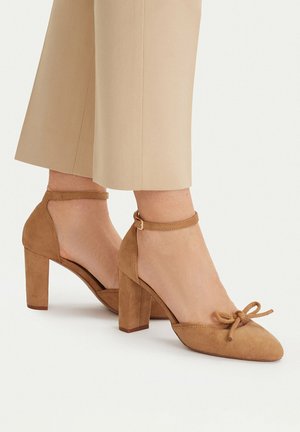 Donna che indossa scarpe beige in camoscio con tacco largo, cinturini alla caviglia e piccolo fiocco, abbinate a pantaloni cropped color beige.