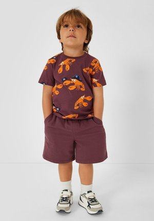 Małe dziecko stoi z rękami w kieszeniach, ubrane w fioletowe spodenki, bordową koszulkę z pomarańczowymi nadrukami homarów, białe skarpetki i trampki.